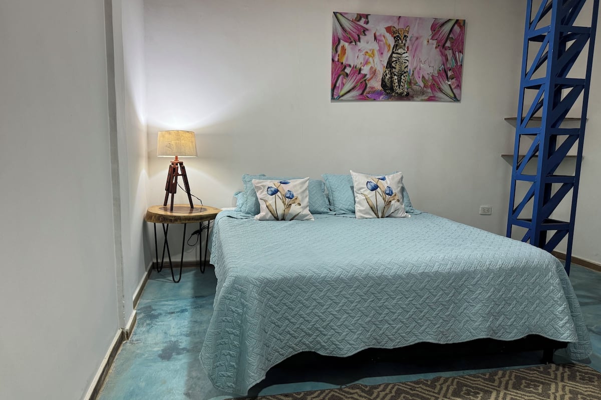 A bedroom with a bed, a nightstand, a lamp, a painting, and a blue metal ladder.