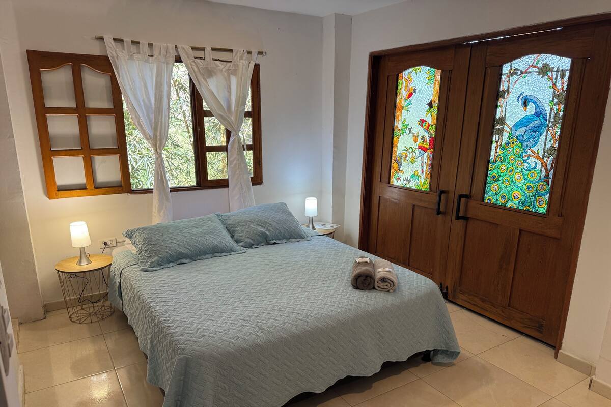 A bedroom with a bed, two bedside lamps, a window with curtains, and a door with stained glass.