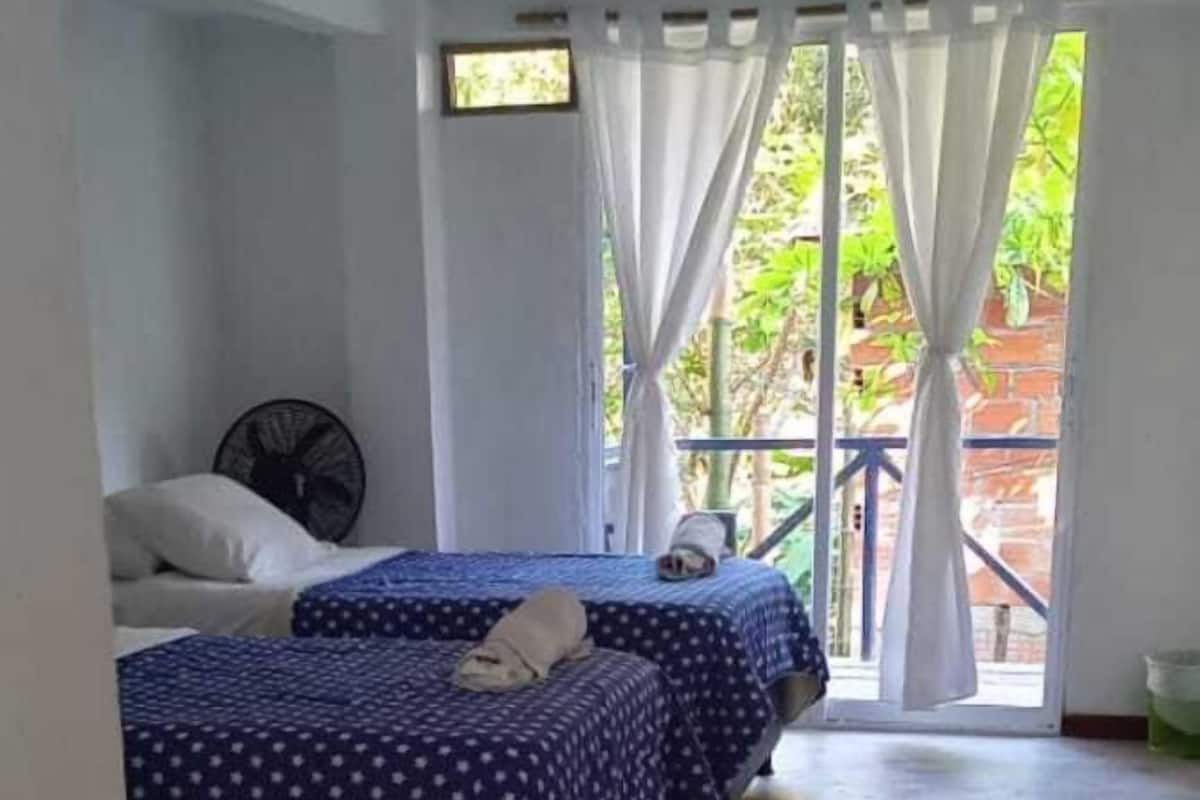 A room with two beds, a fan, and a view of greenery through a window.