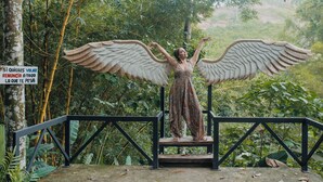 Eine Statue einer Figur mit ausgebreiteten Flügeln, stehend auf einer Plattform mit Geländer, umgeben von üppiger Vegetation.