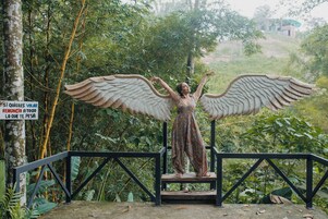 Una estatua de una figura con alas extendidas, sobre un pedestal con barandilla, rodeada de abundante vegetación.