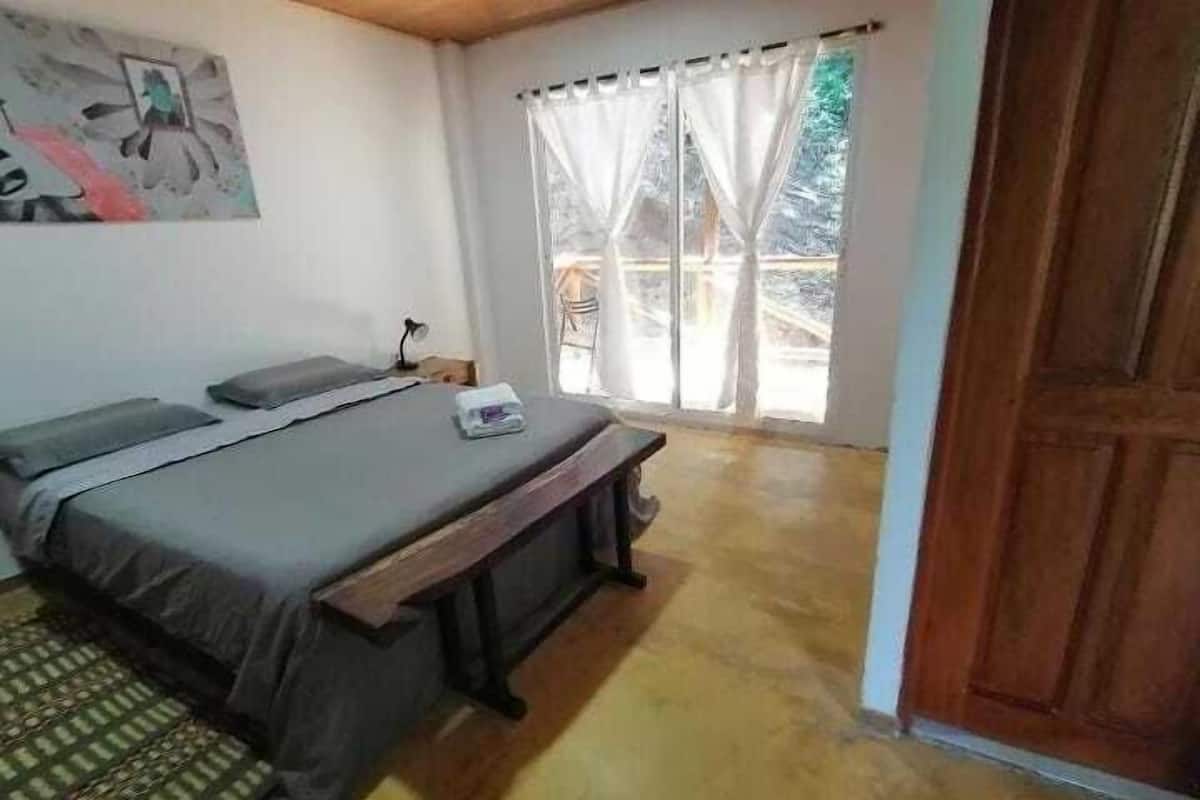 A bedroom with a bed, a bench, a nightstand, a lamp, and a window with curtains.