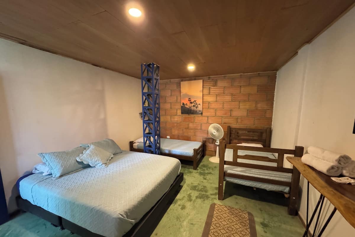 A room with two beds, a wooden chair, a table with towels, a fan, and a brick wall with a painting.