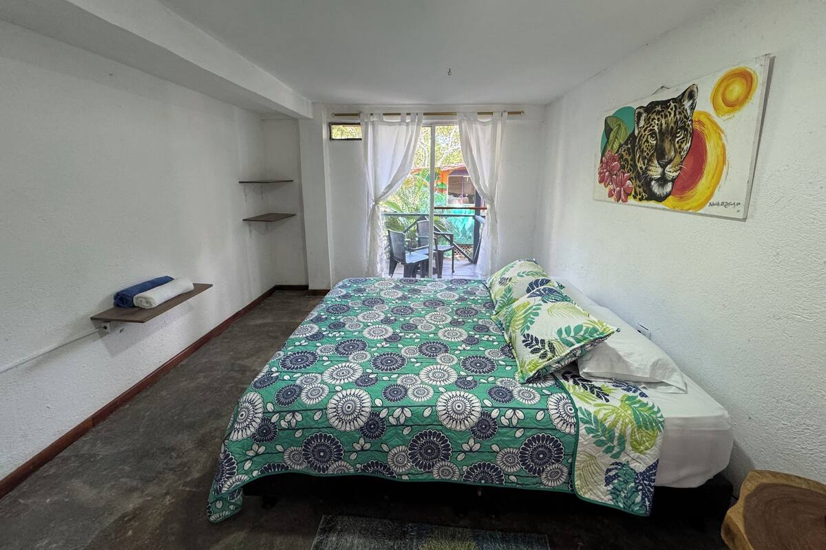 A bedroom with a bed, a painting, a window with curtains, and a view of the outside.