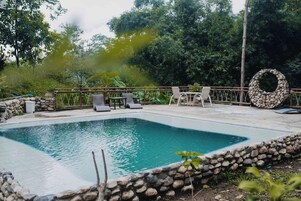 Una piscina rectangular con borde de piedra, rodeada de sillas de descanso y una valla de madera.