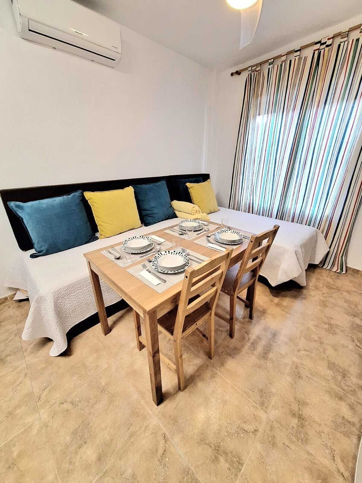 Un coin repas avec une table en bois dressée pour quatre personnes, un canapé agrémenté de coussins bleus et jaunes, et une fenêtre ornée de rideaux à rayures.