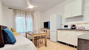 A compact, well-lit room with a bed, a small dining table, a kitchen area with white cabinets, a microwave, and a refrigerator. - Playa Balerma II (Balerma)