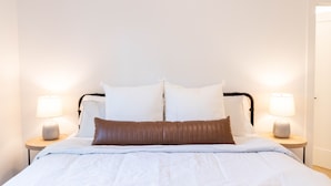 Ein ordentlich bezogenes Bett mit weißer Bettwäsche und zwei braunen Kissen, beides flankiert von zwei Nachttischen mit Lampen.