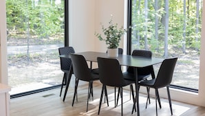 Ein Essbereich mit Tisch und Stühlen, einer Zimmerpflanze und Blick auf einen Wald durch große Fenster.