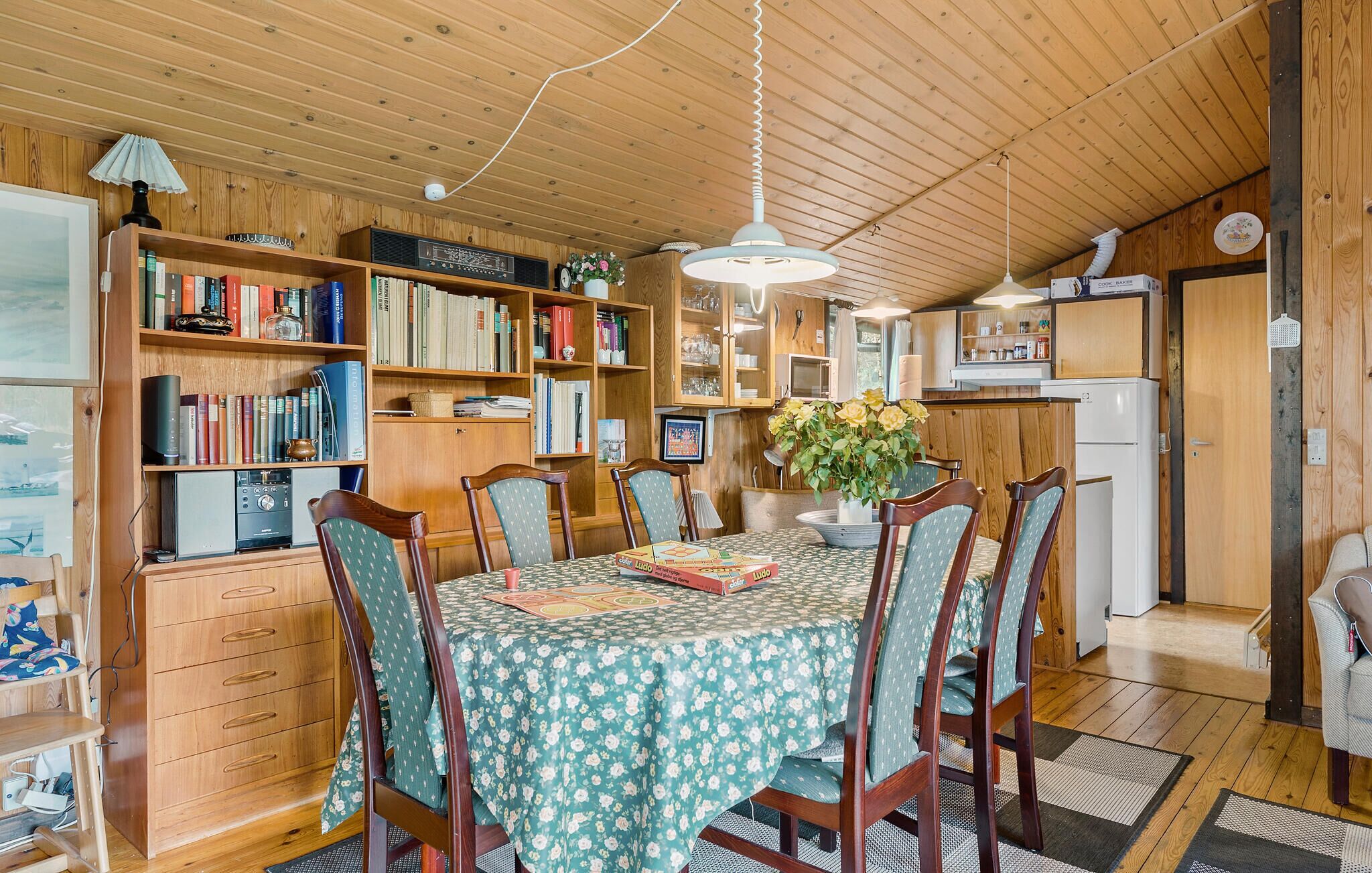 Una stanza accogliente e ben illuminata, con pareti e soffitto in legno, un tavolo da pranzo con tovaglia floreale, sedie e una libreria piena di libri e oggetti decorativi.