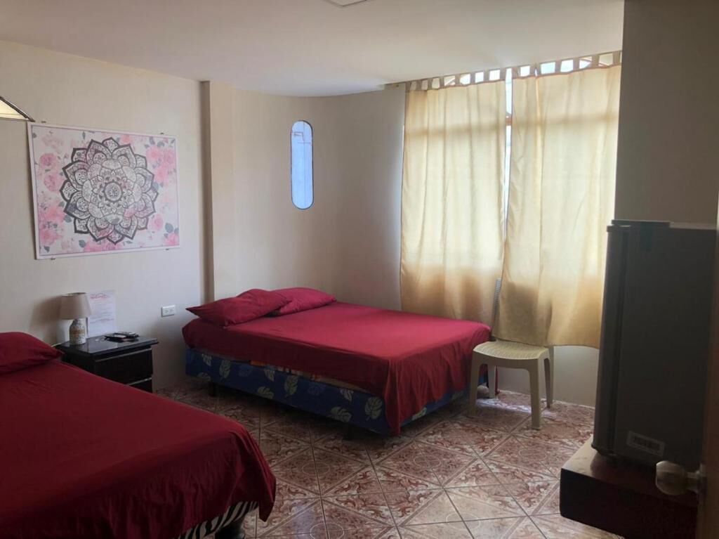 Una habitación con dos camas, una silla, un ventanal con cortinas, un adorno en la pared y un televisor.