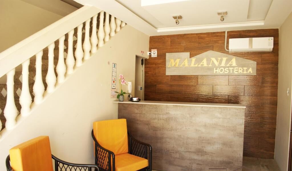 A reception area with a wooden reception desk, a sign reading "MALANIA HOSTERIA", and two chairs with orange cushions.