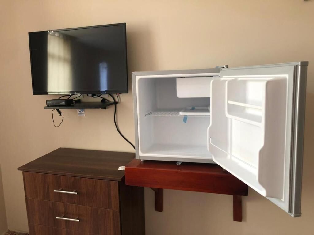 Une pièce de taille modeste, dotée d’un téléviseur à écran plat fixé au mur, d’un meuble en bois et d’un réfrigérateur sans porte.