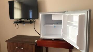 Une pièce de taille modeste, dotée d’un téléviseur à écran plat fixé au mur, d’un meuble en bois et d’un réfrigérateur sans porte.