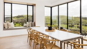 Een eethoek met een lange tafel, houten stoelen en grote ramen met uitzicht op een groen landschap.