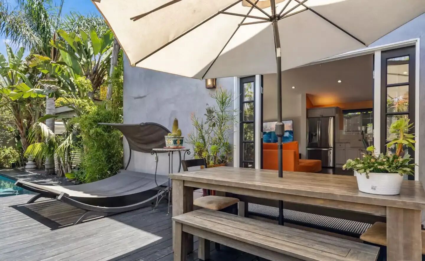 Een modern terras buiten met een houten tafel en bank, omgeven door weelderige begroeiing en een zwembad.