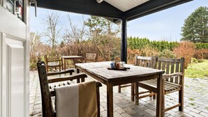 Eine Terrrasse im Freien mit einem Holztisch und Stühlen, einer Bank und Blick auf einen Garten mit Bäumen und Sträuchern.