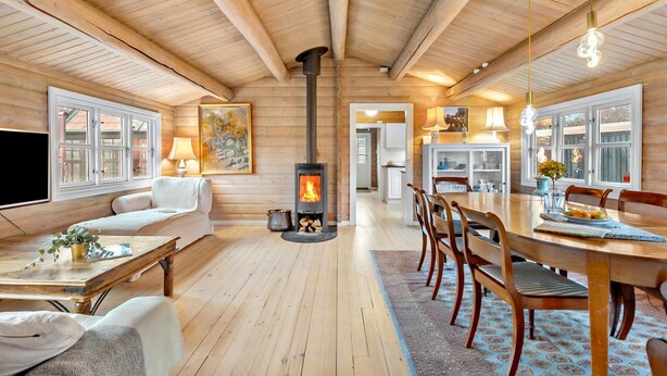 Ein gemütliches Blockhaus-Interieur mit einem zentralen Holzofen, Holzböden und einem Essbereich mit Stühlen und Tisch.
