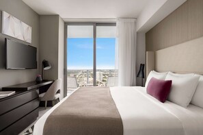 A modern hotel room with a large bed, a desk with a chair, a flat-screen TV, and a city view through large windows.