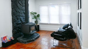 A living room with a stone wall, a black wood-burning stove, a black leather sofa, and a window with blinds. - Private Mountain Creek Getaway! (Vernon Township)