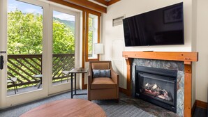 A room with a fireplace, a television mounted on the wall, and a view of greenery outside. - #1315 | STUDIO | The Lodge At Spruce Peak | Big Mountain Views 3rd floor (Stowe)