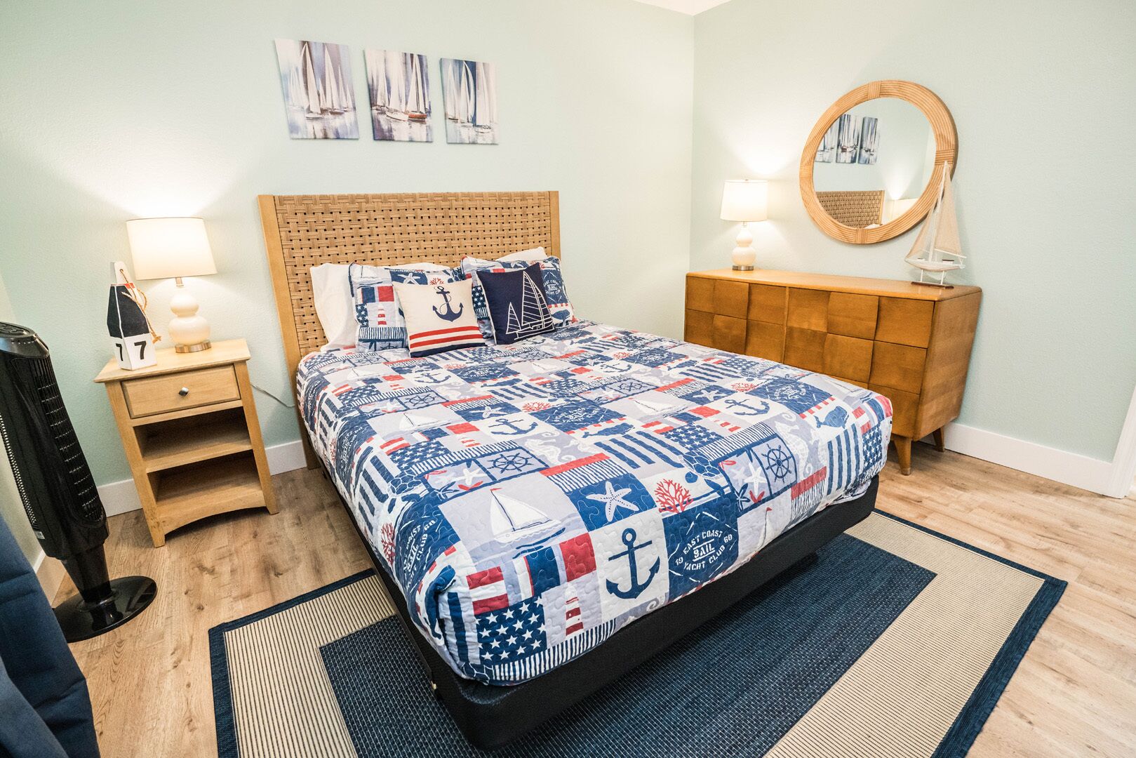 Una camera da letto ordinata con una coperta a tema nautico, una testiera in legno, un comodino con una lampada e un comò con uno specchio.