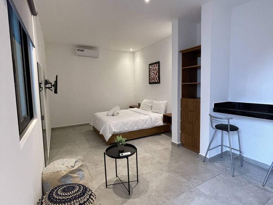 A modern bedroom with a bed, bedside table, and a small stool. There is a desk with a chair and a window.