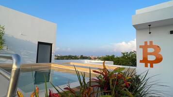 Una piscina en la azotea con una vista despejada del cielo, rodeada de plantas tropicales y el símbolo de Bitcoin en la pared.