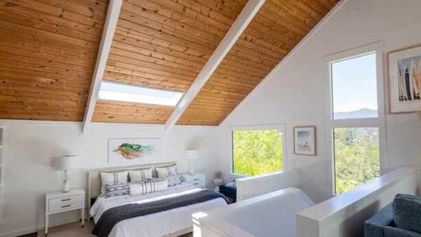 Une chambre avec un plafond en pente, un lit, une table de chevet, une commode, une fenêtre et une vue sur les arbres.