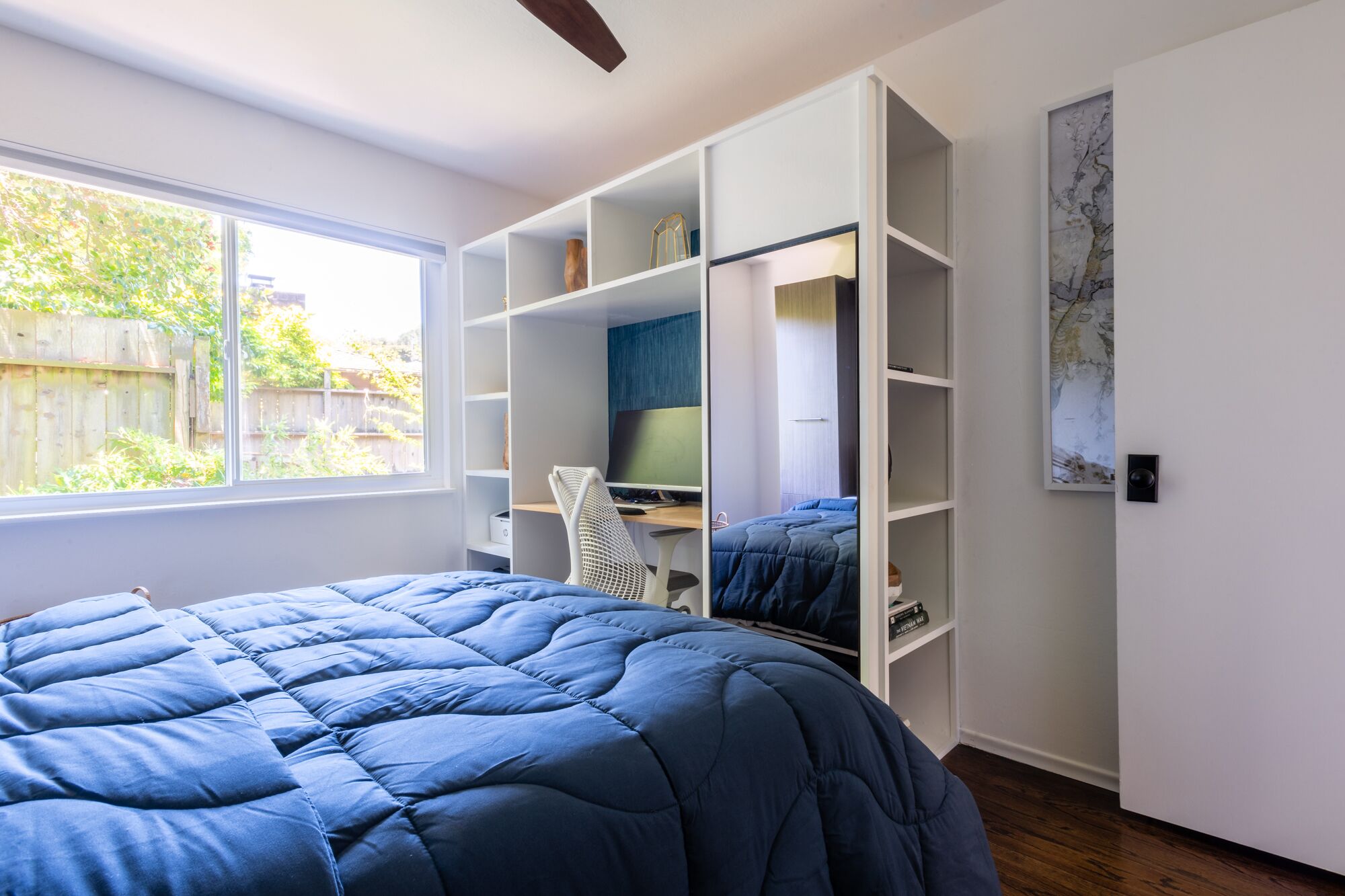 A bedroom with a large bed, a desk, and a computer. There is a window with a view of a fence and trees.