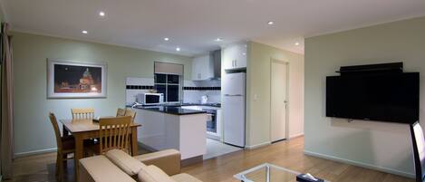 A modern living room with a sofa, a glass coffee table, and a TV. There is a dining area with a table and chairs, and a kitchen with white cabinets and appliances.