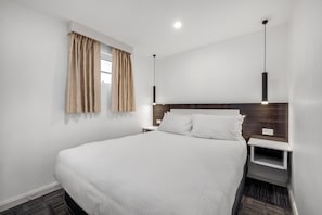 A modern hotel room with a large bed, a bedside table, a nightstand, a window with curtains, and a pendant light. - Buckingham Apartment (Highett)