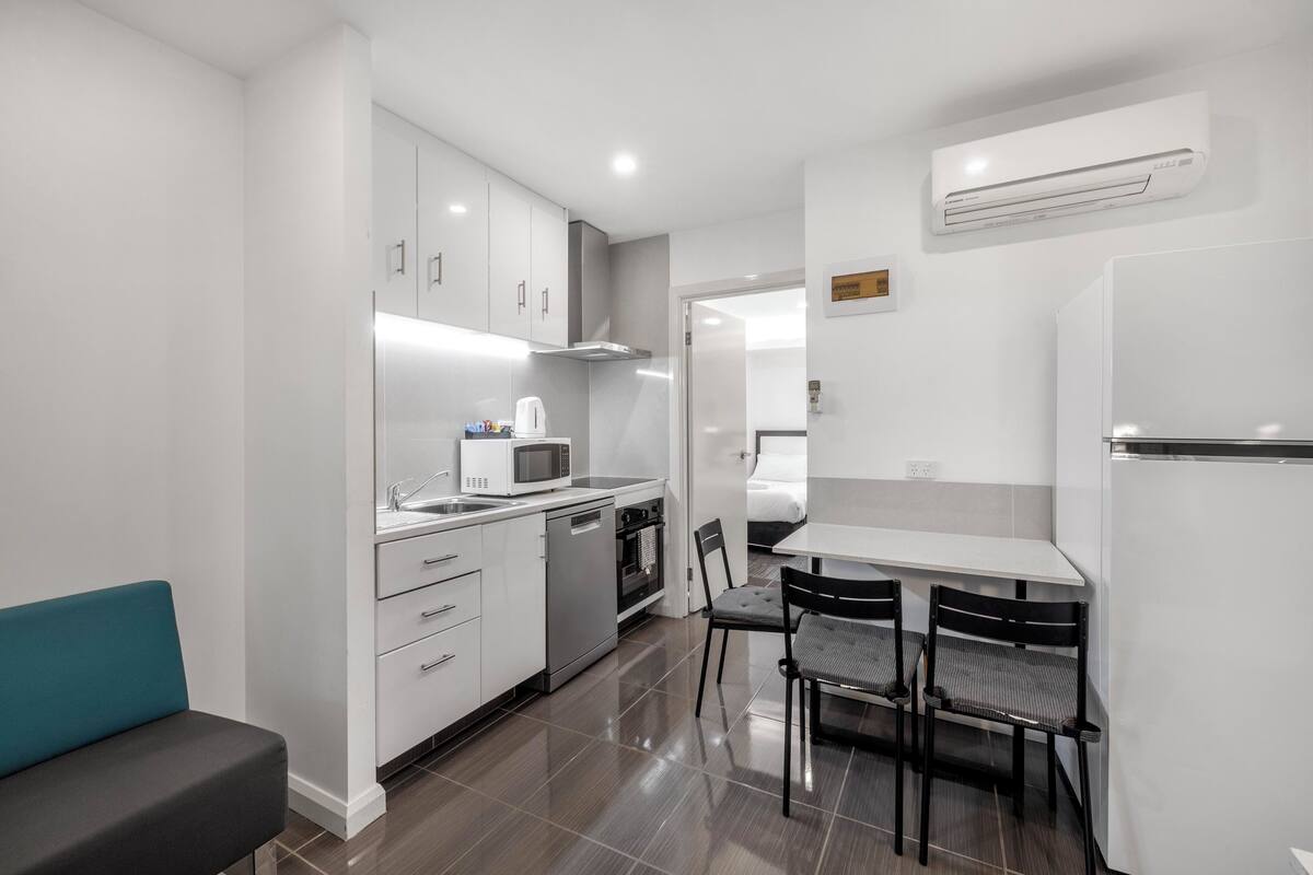 A compact kitchen with white cabinets, a microwave, a dishwasher, and a refrigerator. There is a dining table with two chairs and a bench.