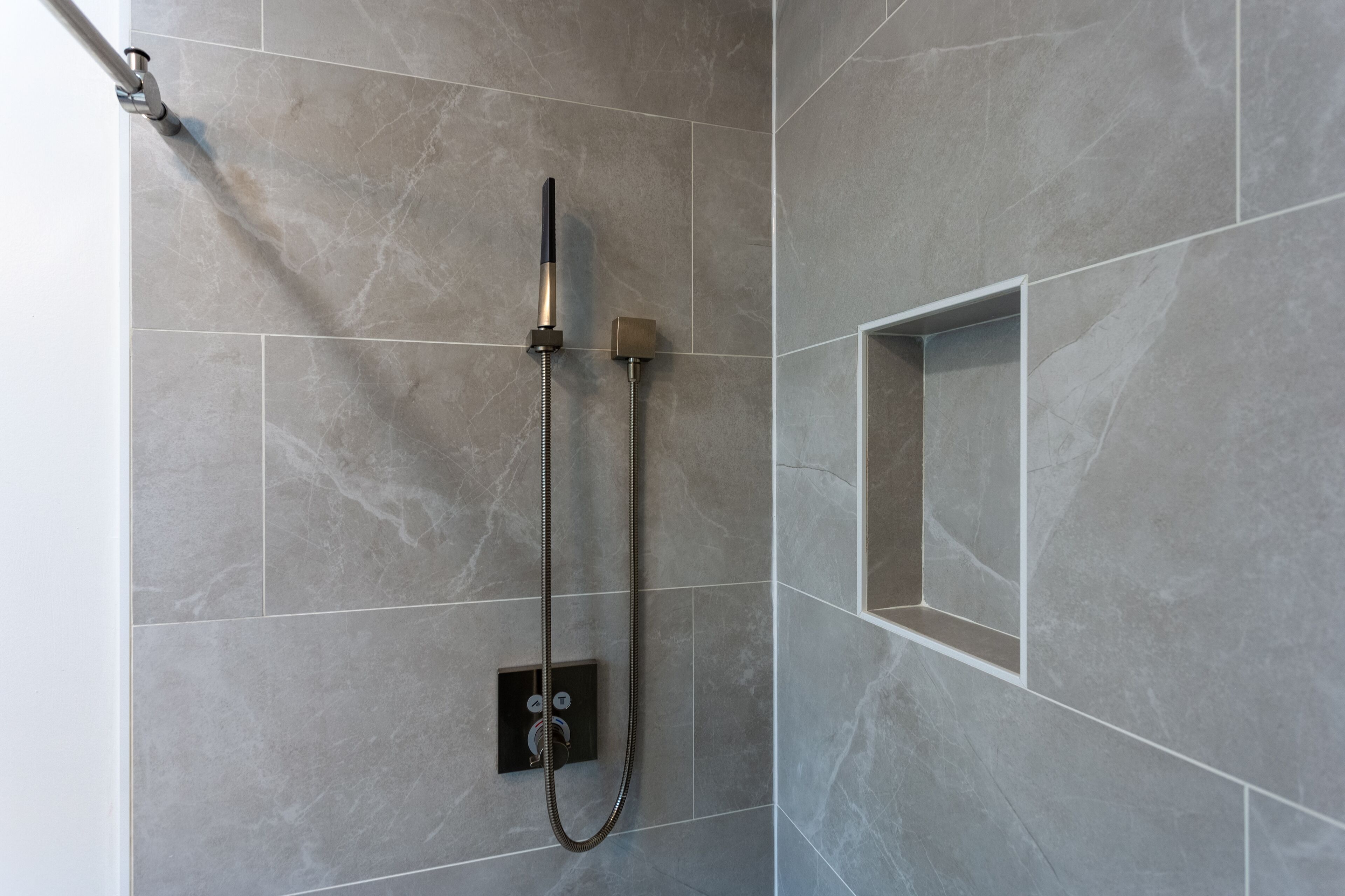 Ducha moderna con cabezal de ducha manual y otro fijo montado en la pared, con un fondo de baldosas de mármol gris.