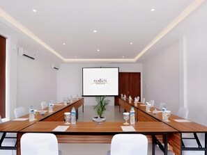 A conference room with a long table, chairs, a projector screen, and a potted plant.