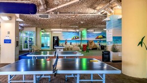 Une pièce équipée de tables de ping-pong, ornée d’une fresque représentant une scène de plage, et sur laquelle est apposée une indication signalant les toilettes.