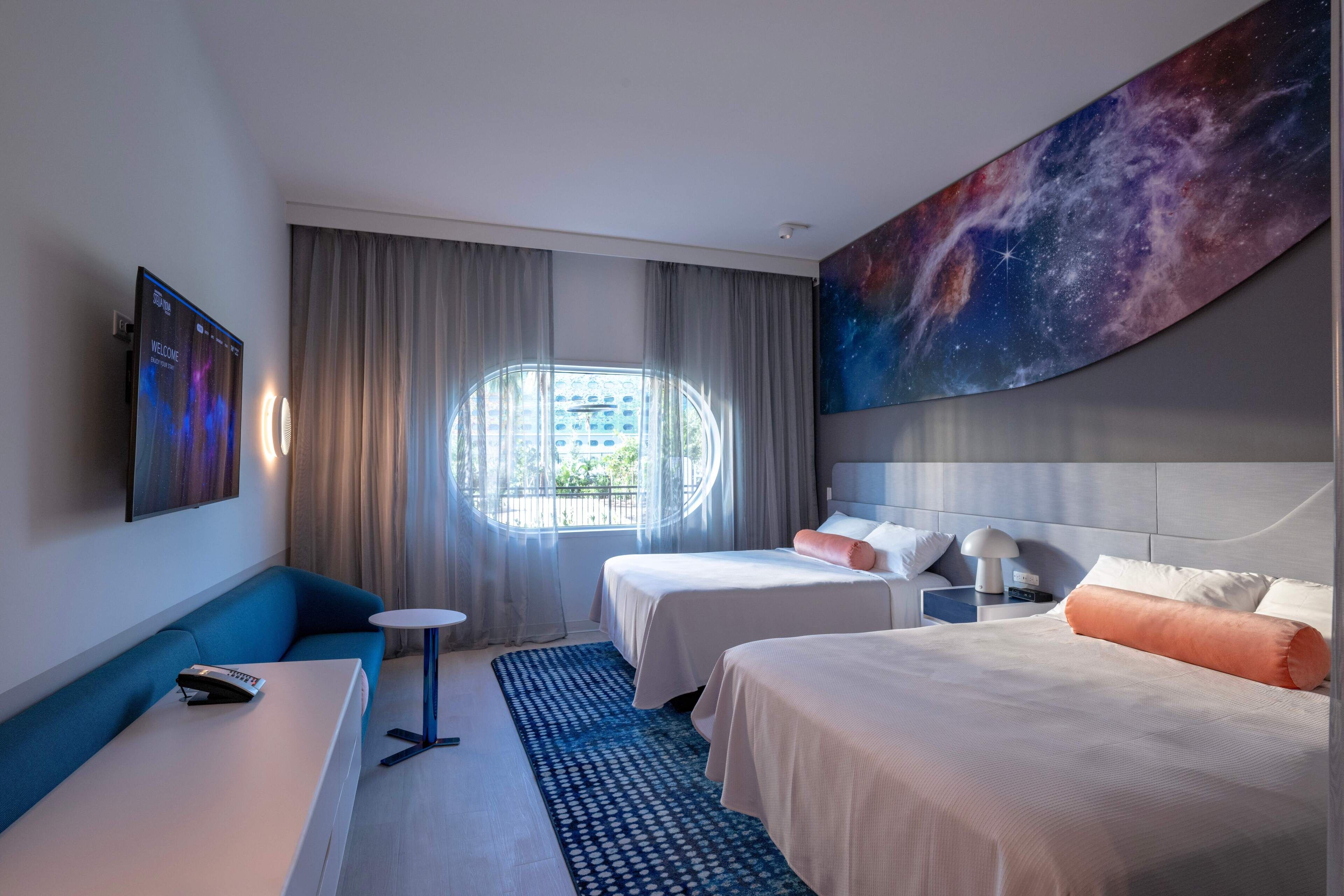 A hotel room with two beds, a sofa, a small table, a TV, and a large painting of a galaxy.
