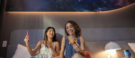 Zwei Personen sitzen auf einem Bett, hinter ihnen eine Wand mit Galaxien-Motiv, und halten ein Smartphone und eine Pizzascheibe.