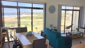 A spacious living area with large windows offering a view of the beach and ocean. There is a dining table with chairs, a blue sofa, and a small coffee table.