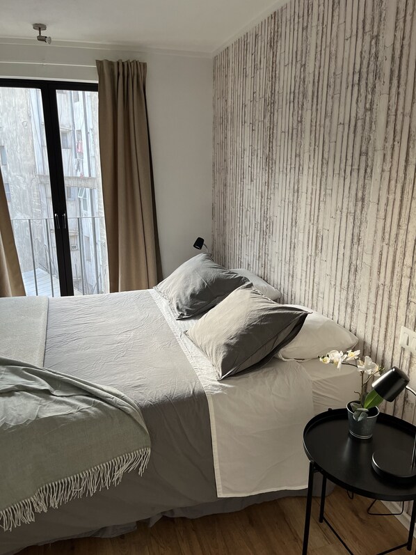 A bedroom with a bed, a bedside table with a lamp, a window with curtains, and a wall with a vertical pattern. - Departamento en Pleno Corazón del Barrio más Bohemio y Entretenido de Santiago (Santiago)