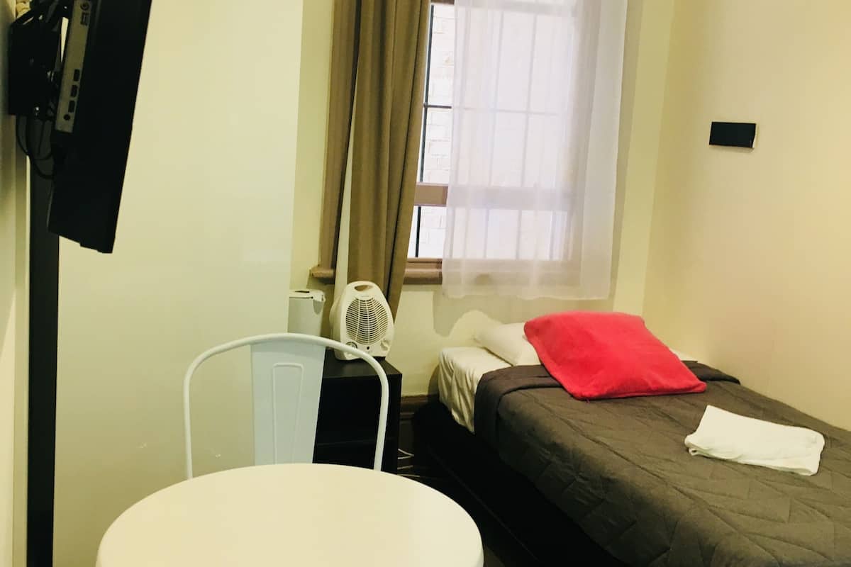 Ein kleines Hotelzimmer mit einem Einzelbett, einem weißen Tisch, einem Stuhl, einem Fernseher und einem Fenster mit Vorhängen.