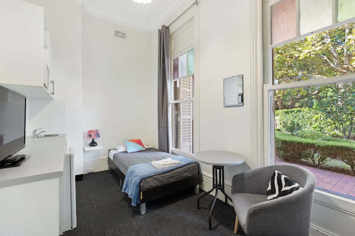 Ein kleines Hotelzimmer mit Bett, einem kleinen Schreibtisch mit Fernseher, einem Sessel und einem Fenster mit Blick auf Grünflächen.