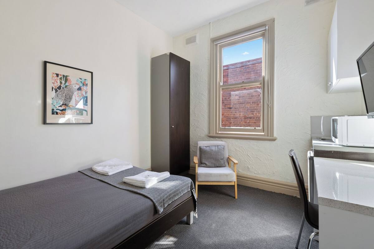 Ein kleines, ordentlich eingerichtetes Schlafzimmer mit einem Einzelbett, einem Sessel, einem Kleiderschrank und einem Fenster mit Blick auf ein Backsteingebäude.