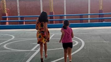 Una cancha de baloncesto al aire libre con valla metálica y pared de ladrillo al fondo.