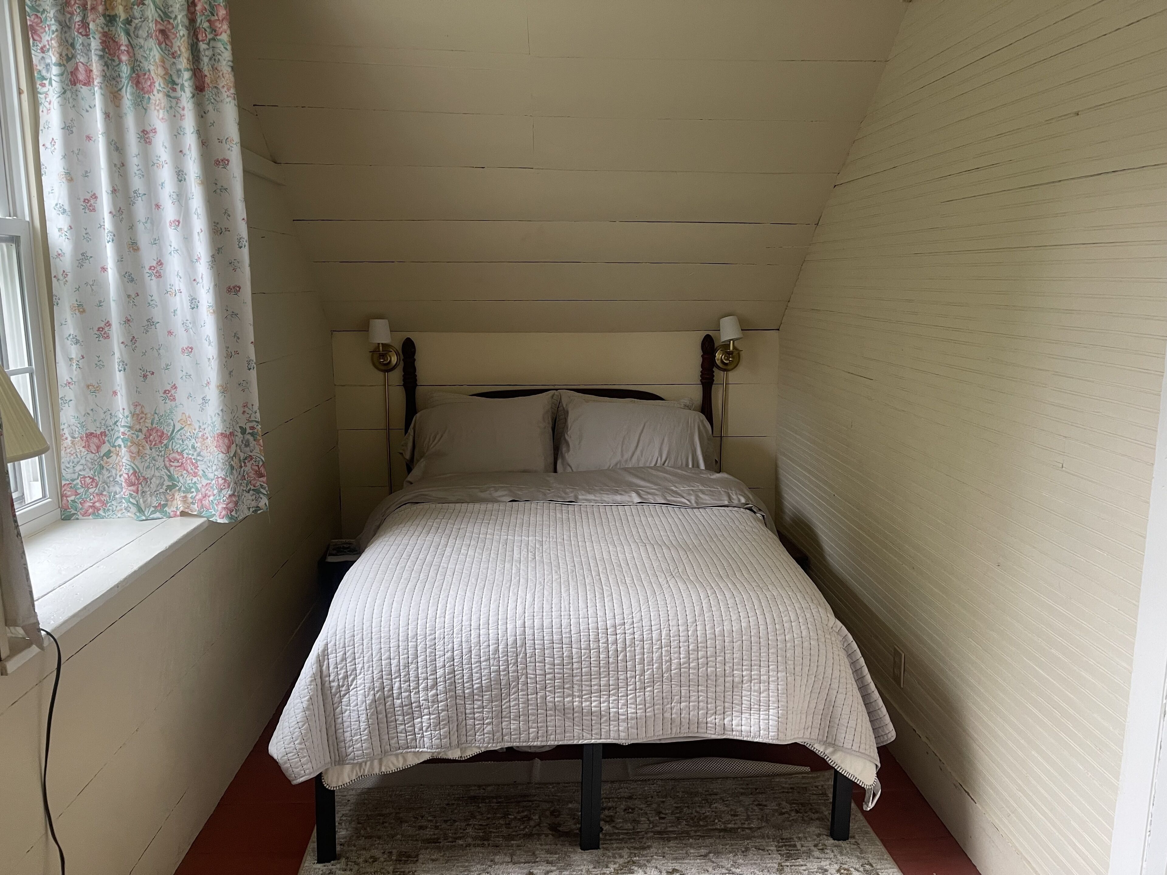 A small, single-bed room with a wooden headboard, a bedside lamp, a floral curtain, and a rug.