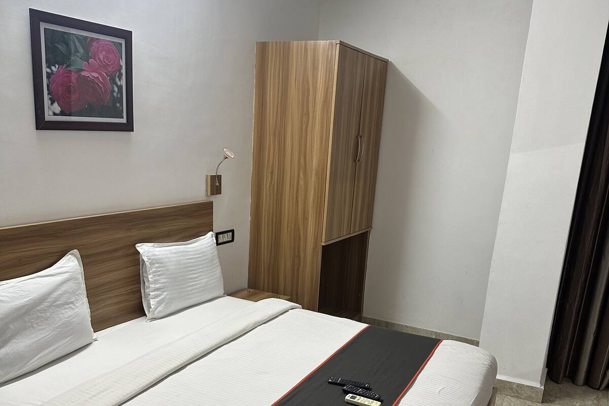 A neatly made bed with white bedding and a black and red accent, a wooden wardrobe, a framed floral picture on the wall, and a small bedside table with a lamp.