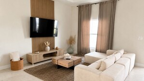 A modern living room with a flat-screen TV, a beige sofa, a wooden coffee table, and a large window with curtains.