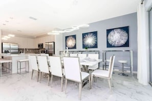 A modern kitchen with white cabinets, stainless steel appliances, and a dining area with white chairs and a table. - 10BD Bella Vida Beautiful Home Near Parks (BV 303) (Kissimmee)