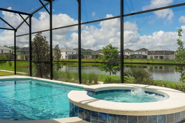 A swimming pool with a hot tub, surrounded by a metal fence, with a view of houses and a body of water in the background.