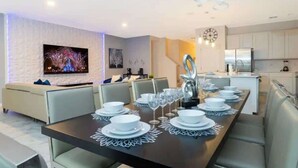 A modern dining area with a long table set for six, a sofa, a TV, and a kitchen in the background.